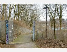 Eingang zu einem Fußweg mit Treppe, flankiert von metallenen Maschendrahtzäunen und Pfosten mit Graffiti. Im Hintergrund kahle Bäume und ein leicht abfallendes Gelände mit Blick auf eine urbane Umgebung.