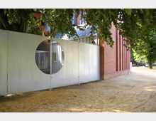 Ansicht einer Backsteinmauer eines Gebäudes mit großen Glasfenstern und einem angrenzenden Metallzaun mit kreisförmiger Öffnung, vor einer sandbedeckten Fläche und umgeben von Bäumen. Im Hintergrund sind parkende Autos und weitere Grünflächen sichtbar.