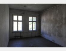 Leerer Raum mit zwei großen, rechteckigen Fenstern mit Rundbögen im oberen Bereich, die Tageslicht hereinlassen. Die Wände sind grau und fleckig, der Boden besteht aus dunklem, abgenutztem Material, und Heizungsrohre verlaufen entlang der Wand unter den Fenstern.