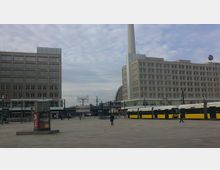 Offener Platz mit zwei großen, quadratischen Gebäuden im Hintergrund, unterbrochen von einer Glaskonstruktion und einem hohen weißen Turm. Im Vordergrund sind gelb-weiße Straßenbahnen sowie eine Litfaßsäule zu erkennen, während wenige Menschen über den Platz gehen.