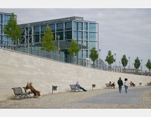 Modernes Stadtbild mit einer gepflasterten Uferpromenade, auf der Bänke stehen, sowie Menschen, die spazieren gehen oder sitzen. Im Hintergrund ein großes Gebäude mit moderner Glasfassade und symmetrischer Architektur, umgeben von einzelnen Bäumen und Laternen.