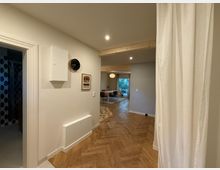 Flur in einem modernen Wohnraum mit hellen Wänden und Holzboden in Fischgrätmuster, an der Wand eine schwarze Wanduhr, ein Wandkasten und ein gerahmtes Bild. Im Hintergrund ein Essbereich mit Tisch, Stühlen und großer Fensterfront, die auf einen Garten blickt.