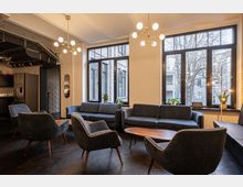 Gemütlicher Gemeinschaftsraum mit modernen Möbeln, darunter dunkelblaue Sessel und Sofas, sowie Holztischen. Große Fenster lassen Tageslicht herein, und goldfarbene Kronleuchter hängen an der Decke, während im Hintergrund eine offene Küche mit einem Kühlschrank zu sehen ist.