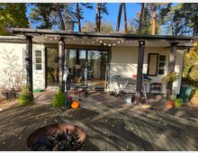 Ein kleines, eingeschossiges Haus mit heller Fassade und einem Flachdach, umgeben von hohen Bäumen. Eine überdachte Holzterrasse mit Dekorationen wie Lichterketten, Pflanzen und Gartenutensilien steht im Vordergrund, während eine Feuerschale auf dem Holzboden platziert ist.