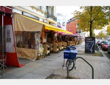 Straßenszene mit Außenterrasse eines indischen Restaurants, das durch gelbe und rote Markisen gekennzeichnet ist. Vor dem Restaurant stehen Tische und beige Stühle, daneben ein Gehweg mit geparkten Fahrrädern und Autos sowie ein Werbeschild mit Mittagsmenü-Angeboten.
