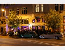 Straßenszene bei Nacht vor einem beleuchteten Gebäude mit großen Fenstern und steinerner Fassade. Vor dem Gebäude stehen viele Menschen, einige Autos sind entlang der Straße geparkt, und Bäume säumen den Gehweg.