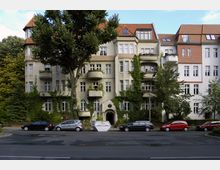 Mehrstöckiges Wohngebäude mit historischer Fassade, Balkonen und rankender Begrünung, flankiert von Bäumen. Autos parken vor dem Gebäude entlang einer Straße.
