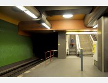 U-Bahn-Station mit einem Gleisbereich links, einer Wand mit grünen Kacheln und einer Rolltreppe sowie Treppenaufgang zur Ausgängebene rechts. Im Vordergrund ein Ticketautomat und Geländer.