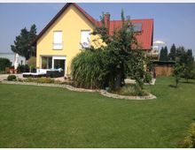 Ein gelbes Einfamilienhaus mit rotem Satteldach, umgeben von einem gepflegten Garten mit Rasenflächen, Bäumen und Beeten. Auf der Terrasse stehen Gartenmöbel und im Hintergrund sind weitere Bäume und ein Holzanbau sichtbar.