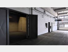 Leerer Innenraum einer Industriehalle mit weiß gestrichenen Betonwänden, sichtbaren Rohrleitungen an der Decke und großen Fenstern auf der rechten Seite. Links im Bild befinden sich eine geöffnete Metalltür und eine kleinere Nische.