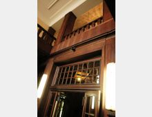 Holzvertäfelter Innenraum mit einem Eingang, der aus verglasten Holztüren und einem dekorativen Oberlicht mit kleinen Glasquadraten besteht. Im oberen Bereich ist eine hölzerne Balustrade sichtbar, die eine Zwischengalerie begrenzt.