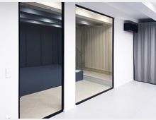 Minimalistischer Innenraum mit weißen Wänden, großen Glaswänden in schwarzem Rahmen und hellgrauem Vorhang. Im Hintergrund ist ein Treppenbereich mit Sichtbeton und leicht industriellem Design sichtbar.