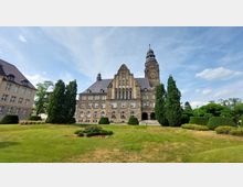 Historisches Gebäude aus Sandstein mit zahlreichen Fenstern, einem hohen zentralen Giebel und einem Turm mit Uhr. Vor dem Gebäude befindet sich eine gepflegte Rasenfläche mit Blumenbeeten und hohen Bäumen.
