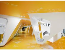 Futuristisch gestalteter Innenraum mit schrägen weißen und gelben Wänden sowie einem gelben Boden, der mit weißen Illustrationen verziert ist. Große Öffnungen in den Wänden und Fenster im Hintergrund lassen Tageslicht einfallen.