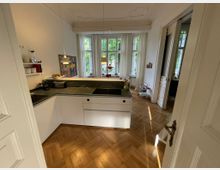 Moderne Küche in einem Altbau mit Fischgrät-Holzboden, weißen Schränken und einer Arbeitsplatte aus dunklem Stein. Große Fenster mit weißen Vorhängen lassen viel Tageslicht herein; im Hintergrund ist ein Essbereich sichtbar.