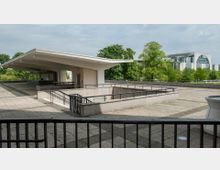 Offener Dachbereich mit modernem Flachdach-Pavillon und umlaufendem Geländer, umgeben von einer gepflasterten Terrasse. Im Hintergrund sind Bäume und ein modernes Regierungsgebäude mit geschwungener Glasfassade sichtbar.