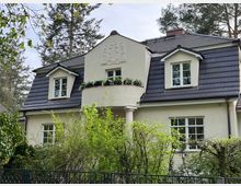 Zweistöckiges Wohnhaus mit cremefarbener Fassade und dunkel gedecktem Mansarddach, mit zwei Gaubenfenstern und einem mittigen, halbrunden Balkon mit Blumenkasten. Im Vordergrund stehen dichte Sträucher und ein Zaun, das Haus liegt in einer von hohen Bäumen umgebenen Garten- oder Waldumgebung.