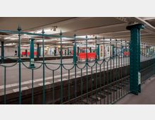 U-Bahn-Station "Wittenbergplatz" in Berlin mit beleuchtetem Bahnsteig, grünen Stützpfeilern, roten Sitzbänken und Werbeplakaten an den Wänden. Im Vordergrund ist ein dunkelgrünes Zaungitter zu sehen.