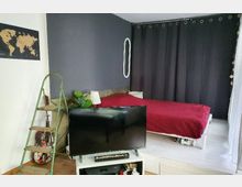 Schlafzimmer mit dunkler Akzentwand, einem Bett mit roter Bettdecke, dunklen Vorhängen und einem modernen Fernseher auf einem weißen TV-Regal im Vordergrund. Links steht eine grüne Leiter mit Dekorationen, darunter eine Gitarre, und an der Wand hängt eine Weltkarte.