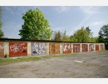 Reihe von einfachen Garagen mit bemalten Holztoren, bedeckt mit Graffiti, vor einer grünen Baumlandschaft und einem offenen, unbefestigten Boden.