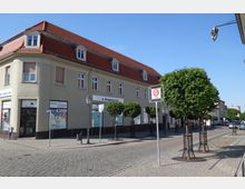 Ecke eines zweigeschossigen Gebäudes mit cremefarbener Fassade, roten Dachziegeln und kleinen Dachfenstern, im Erdgeschoss befindet sich eine Drogerie. Vor dem Gebäude stehen gepflasterte Gehwege, junge Bäume und ein Schild für eine Tempo-20-Zone, im Hintergrund eine ruhige Straße mit parkenden Autos.