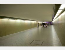Ein unterirdischer Fußgängertunnel mit gelblichen Kacheln an den Wänden und Deckenbeleuchtung. Im Hintergrund läuft eine kleine Gruppe von Menschen Richtung Ausgang, dessen Bereich leicht lila beleuchtet ist.