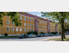 Wohngebäude mit gelber Fassade und roten Akzentfeldern, dreigeschossig und mit zahlreichen weißen Fenstern, entlang einer Kopfsteinpflasterstraße. Im Vordergrund parkende Autos und Bäume auf beiden Straßenseiten.