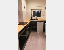 Kleine Küchenzeile mit weiß gefliesten Wänden, einer Arbeitsplatte aus hellem Holz, einem schwarzen Spülbecken und Geräten wie einem Backofen, einem Kochfeld und einer Spülmaschine. Ein Fenster befindet sich an der Stirnseite, und der Boden ist mit hellem Laminat gestaltet.