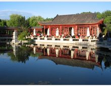 Traditioneller chinesischer Pavillon mit geschwungenem, grauem Ziegeldach und roten Holzfassaden mit dekorativen Gitterfenstern an einem Teich. Im Vordergrund spiegeln sich Gebäude, Sonnenschirme und eine kleine steinerne Pagode im ruhigen Wasser, umgeben von dichtem Grün.