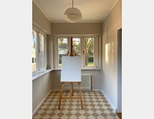 Heller Raum mit cremefarbenen Wänden und kariertem Boden in Beige- und Grautönen, ausgestattet mit einem Holzstaffelei-Ständer und leerer Leinwand vor einem Heizkörper unter drei Fenstern. Außerhalb der Fenster ist ein Baum und eine Gartenlandschaft sichtbar.