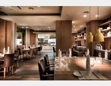 Modernes Restaurant mit eleganter Holzeinrichtung, gedeckten Tischen und Ledersesseln. Im Hintergrund eine offene Küche und Regale mit Dekorationsartikeln, sowie goldene Hängelampen über einem Tisch im Vordergrund.