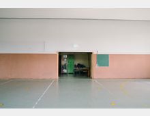 Innenansicht einer Turnhalle mit beige verkleideten Wänden und einem grünen Boden. Im Hintergrund ist eine offene Tür zu einem Lagerraum mit grünen Schränken, zusammengelegten Matten und Stühlen sichtbar; daneben hängt eine grüne Schreibtafel an der Wand.