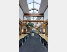 Ein Innenraum einer Bibliothek oder eines Lernraums mit hohen Holzbalken, großen Dachfenstern und symmetrisch angeordneten Tischen sowie Stühlen. In der Mitte steht ein großer Pflanzbehälter mit Grünpflanzen, die den Raum beleben.