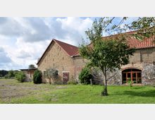 Ein großes Bauernhaus aus Backstein mit rotem Ziegeldach, teilweise mit Bruchsteinfundament. Im Vordergrund befindet sich eine grüne Wiese mit Bäumen, während der Himmel von Wolken bedeckt ist.