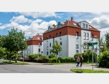 Mehrstöckige Wohngebäude mit weißer Fassade und roten Ziegeldächern stehen in einer ruhigen Wohnstraße mit Fußwegen, Bäumen und Straßenschildern. Im Vordergrund laufen zwei Personen auf einem Gehweg, umgeben von gepflegten Grünflächen.