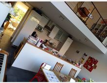 Moderne Wohnküche mit weißer Einbauküche, Holzarbeitsplatte und dekorativen Details. Im Vordergrund ein Esstisch mit Holzstühlen und Blumen, im Hintergrund ein offener Durchgang zu einem weiteren Raum sowie eine offene Galerie mit Treppenzugang.