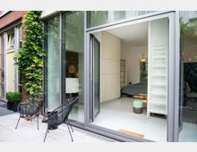 Ground-level patio of a modern brick-and-glass apartment with large sliding doors opening into a minimalist bedroom. The terrace has two black wire chairs and potted greenery, with climbing vines on the exterior wall and a white ladder-style shelving unit visible inside.