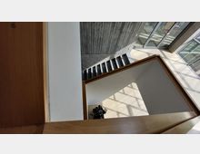 Innenansicht eines modernen Gebäudes mit Beton- und Glasarchitektur. Zu sehen ist eine Treppe neben einer Wand aus Sichtbeton, helle Bodenflächen mit Licht- und Schattenmustern sowie ein Bereich mit Glastüren im Hintergrund.