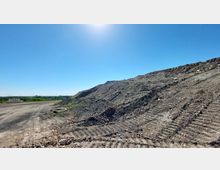 Eine aufgeschüttete Erdhalde mit deutlich sichtbaren Reifenspuren auf der Oberfläche, umgeben von einer kargen Landschaft unter einem klaren, sonnigen Himmel. Im Hintergrund sind Bäume sowie eine Absperrung am Rand eines Weges erkennbar.