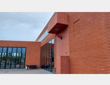 Moderne Backsteinfassade eines Gebäudes mit großen, vertikal ausgerichteten Glasfenstern. Über einem Eingang ist ein rechteckiger Vorsprung zu sehen, daneben hängt eine rote Wanddekoration in Form eines Drachenkopfes.