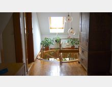 Ein kleiner, heller Wohnraum mit einer Dachfensteröffnung, hellem Holzfußboden und einem runden Treppengeländer aus Holz, das nach unten führt. Topfpflanzen und kugelförmige Hängeleuchten zieren den Bereich, und im Vordergrund stehen Holzschränke und Türen.