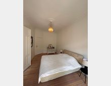 Schlicht eingerichtetes Schlafzimmer mit Holzboden, einem Bett mit Holzrahmen, einer kleinen Kommode samt Spiegel und einer Hängelampe mit goldfarbenen Details. Die Wände und Decke sind weiß, mit dezenten Stuckverzierungen an der Decke.