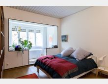 Schlafzimmer in modernem Design mit Holzboden, einem Doppelbett mit blauer Bettwäsche und roter Decke, daneben ein Nachttisch mit Lampe. Im Hintergrund öffnet sich ein Bereich mit großen Fenstern und einer freistehenden Badewanne, umgeben von Pflanzen.