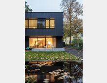 Modernes zweistöckiges Wohnhaus mit dunkler Holzfassade und großen Fenstern, die den Innenraum beleuchten. Im Vordergrund ein kleiner Teich, eine Grünfläche mit Herbstlaub, sowie ein umliegender Garten mit Bäumen und benachbarten Wohnhäusern.