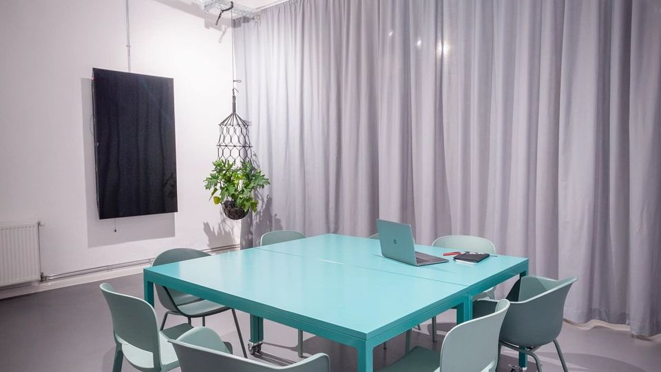 Moderner Besprechungsraum mit türkisfarbenem Tisch auf Rollen und mehreren passenden Stühlen auf grauem Boden. An der Decke hängen schallabsorbierende Paneele und Strahler, an der Wand befinden sich ein schwarzes Paneel sowie eine große graue Vorhangfront und eine hängende Pflanze.