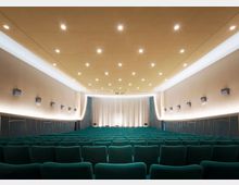 Großer Kinosaal mit Reihen aus grünen Polstersitzen, einer Bühne mit zugezogenen, weißen Vorhängen und einer goldfarbenen Decke mit zahlreichen kleinen Deckenleuchten. Die Wände sind beige, mit integrierten Lautsprechern und indirekter Beleuchtung.