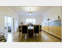 Esszimmer mit Holzfußboden, einem großen rechteckigen Tisch und acht Stühlen mit dunklen Polstern. Rechts steht ein lange Holzvitrine mit teils satinierten Glastüren; eine Schiebetür führt in ein angrenzendes Wohnzimmer mit Sofa.