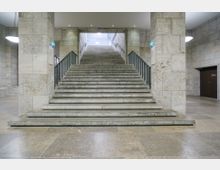 Breite Steintreppe in einem Gebäude mit hellen Natursteinwänden, flankiert von Metallgeländern. Die Treppe führt in einen beleuchteten oberen Bereich, während an den Seiten massive Wände und eine Tür sichtbar sind.