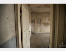 Ein leerer, heruntergekommener Raum mit schmutzigen, fleckigen Wänden und einfachem Betonboden. Im Vordergrund ist eine geöffnete, gealterte Holztür sichtbar.