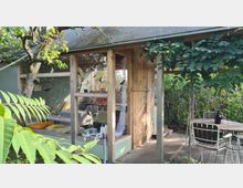 Ein kleines, rustikales Gartenhäuschen mit großen Fenstern, einer Holztür und einem umlaufenden Holzdeck, umgeben von üppigen Pflanzen und einem Obstbaum. Vor dem Häuschen steht ein runder Holztisch mit Metallstühlen, daneben rankt grünes Laub an einer Pergola.